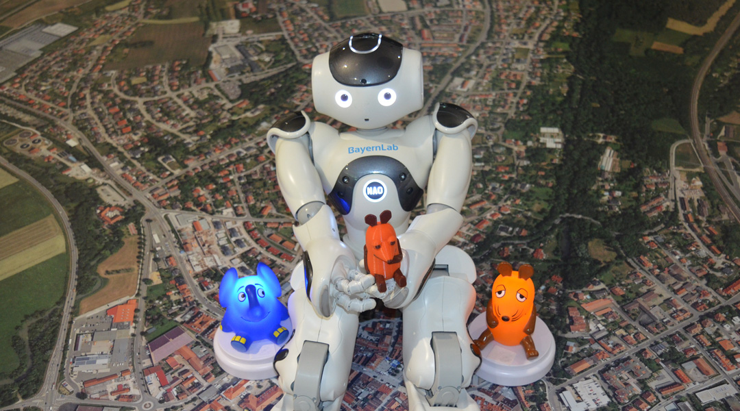 Roboter Nao sitzt auf einem Luftbild und hält die Maus in der Hand, links und rechts der Elefant und die Maus von innen beleuchtet